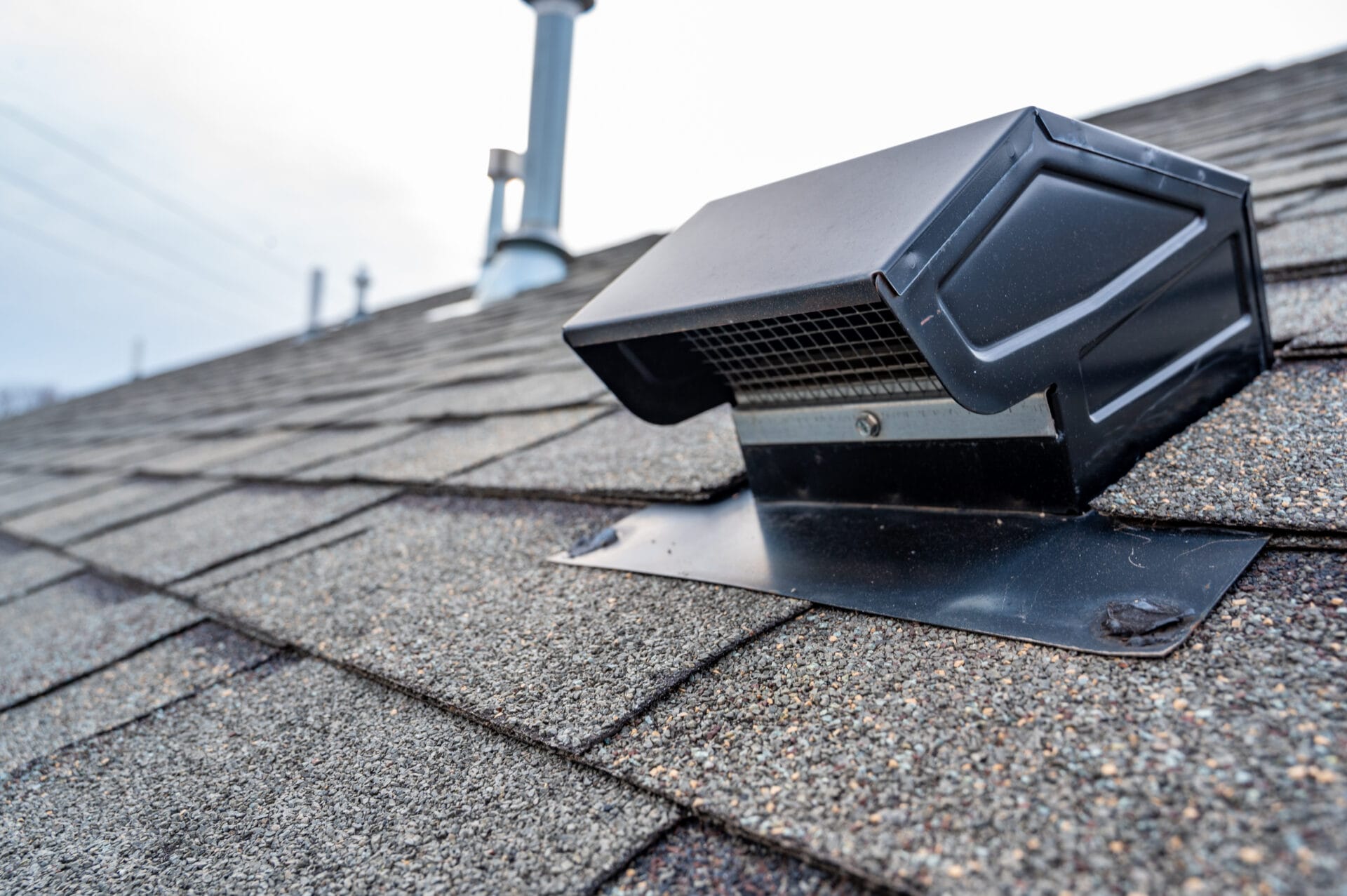 Close-up of a black roof vent installed on asphalt shingles, with pipes and vents visible in the background against a cloudy sky—a helpful image when considering questions to ask roofers about proper ventilation.