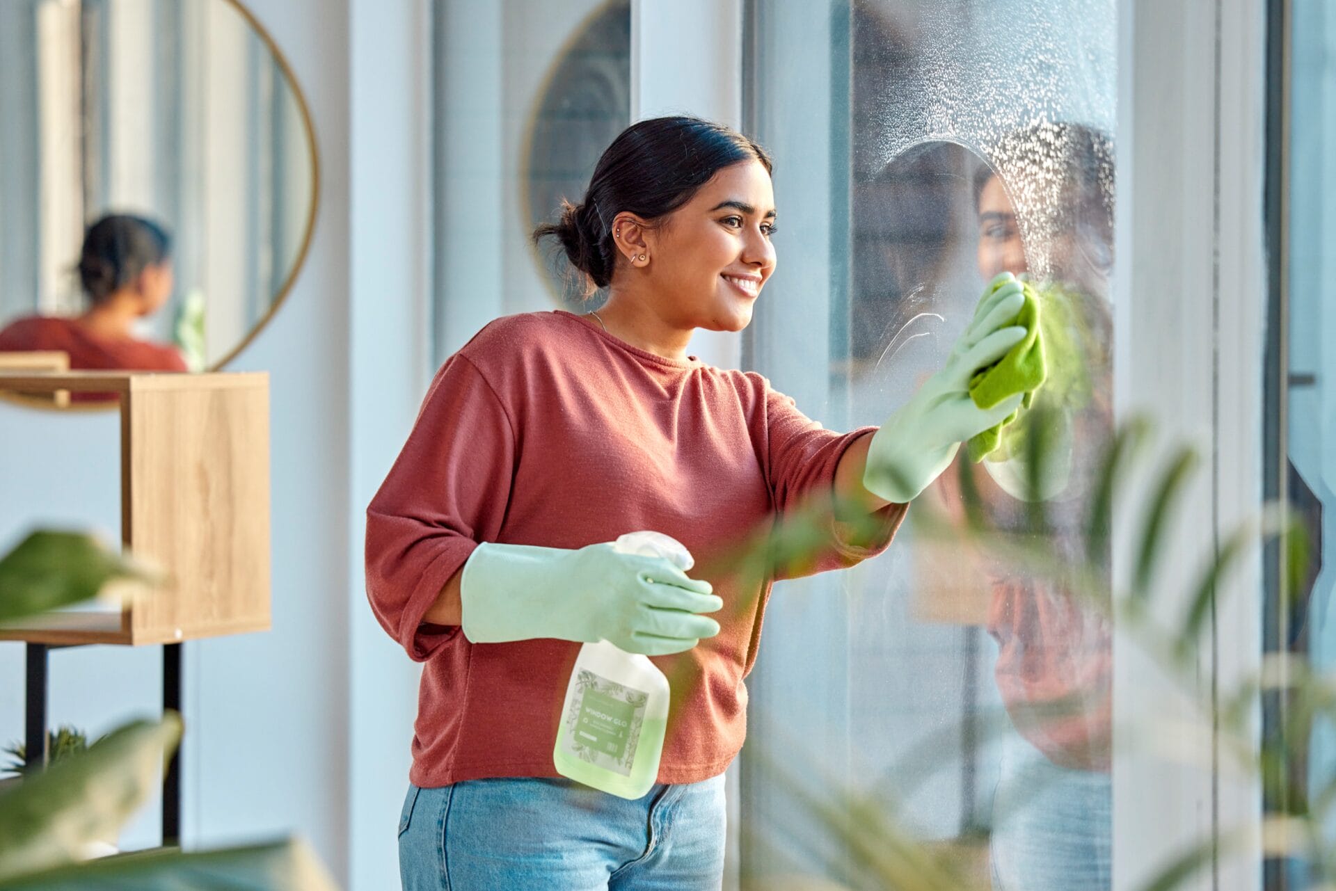A woman wearing gloves cleans a window with a spray bottle and cloth, standing indoors near a round wall mirror and leafy plants, putting DIY window maintenance tips into practice.
