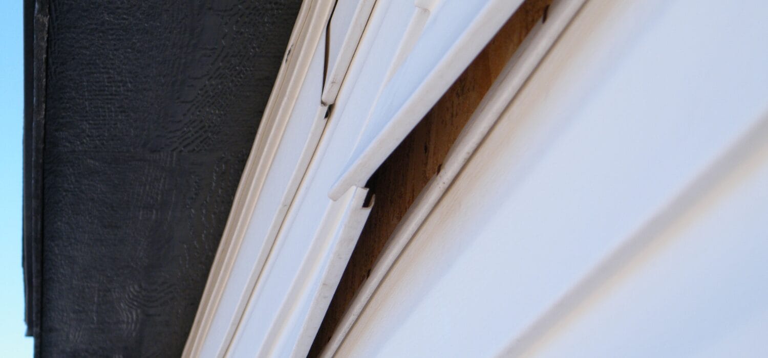 Close-up view of white house siding panels detached and warping outward, revealing exposed wood beneath, next to a dark vertical wall.