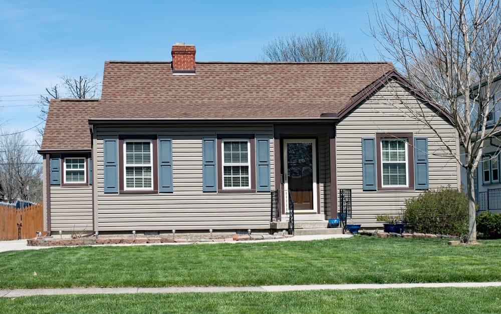 Single-story house with tan siding, brown roof, blue shutters, a small front porch, and a well-maintained lawn under a clear blue sky.